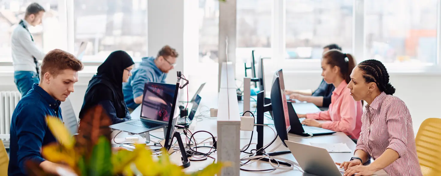 An einem großen tisch sitzen sich viele Mitarbeitende mit ihren PCs gegenüber und arbeiten konzentriert.